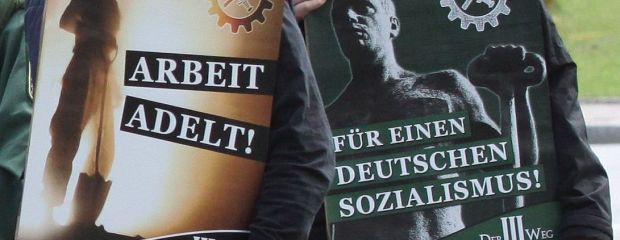 Auf dem Bild sind zwei Aktivisten der Partei Der Dritte Weg zu sehen, die Kundgebungsplakate mit den Aufschriften "Arbeit adelt" und "Für einen deutschen Sozialismus!" halten. 