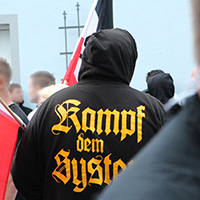 Auf dem Foto ist ein Teilnehmer einer rechtsextremistischen Kundgebung zu sehen, der eine Jacke mit der Aufschrift "Gegen das System" trägt.
